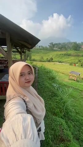 A woman in a hijab takes a selfie, waving at the camera. The background features lush green rice paddies and a hillside dotted with trees and buildings. The sky is blue with some white clouds. The woman is wearing a light-colored outfit and a purse with a chain strap. She appears to be standing near a wooden structure, possibly a gazebo or covered porch. The overall setting is a rural, agricultural landscape.