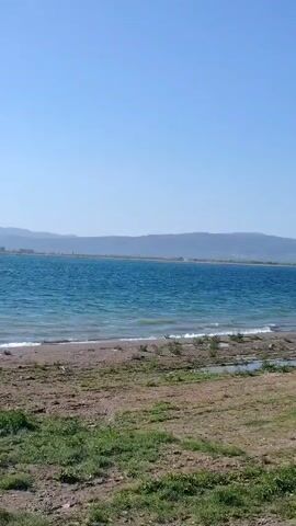 This video captures a wide, serene shot of Iznik Lake in Iznik, Turkey, on a bright, sunny, and windy day.

The foreground reveals an unpaved shoreline composed of a mix of sand and earth, dotted with sparse patches of green grass and dry vegetation. Small waves gently break along the water's edge, forming a narrow band of white foam.

Beyond the immediate shore, the vast expanse of Iznik Lake stretches out. The water is a vibrant blue, agitated by the wind, resulting in a choppy surface with numerous small waves. As the camera subtly pans to the right, the sun's reflection intensifies on the water, creating a dazzling, shimmering spectacle that covers a significant portion of the lake's surface. In the mid-section of the video, a few small, dark birds can be observed wading or standing on the wet sand near the waterline.

In the background, a long, indistinct chain of mountains or hills borders the far side of the lake, appearing somewhat hazy under the clear, light blue sky. The distinct sound of wind is audible throughout the recording, underscoring the breezy conditions. No text is visible in the video.
