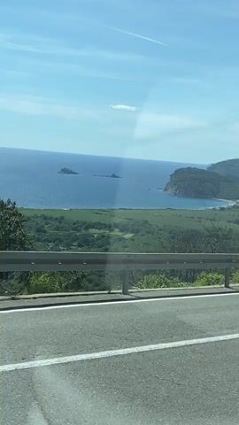 The video shows a view from a moving vehicle on a sunny day. In the foreground, there is an asphalt road with white lane markings. A silver guardrail runs along the side of the road. Beyond the guardrail, there is a green, grassy slope with some sparse trees. In the mid-ground, a body of water, likely the sea, is visible under a bright blue sky with a few white clouds and a faint contrail. The sea is dotted with a few small rocky islands and meets a coastline with cliffs and a sandy beach. Some buildings are visible nestled along the coast. In the first few seconds, a blue car with a roof rack passes by in the opposite lane. A circular black sign with some white text is briefly visible on the right side of the road in the latter half of the video.