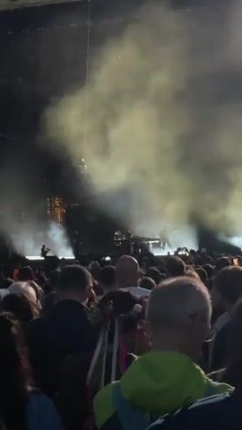 The video shows a crowded outdoor concert at night. A band is performing on a stage illuminated by bright lights and thick smoke. The audience is densely packed, with many people holding up their phones to record. The sign "OPEN'ER FESTIVAL" is visible at the top of the stage.