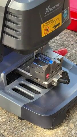 This is a close-up, eye-level shot of a Xhorse automatic key cutting machine in operation. The machine is dark gray and black with a prominent yellow "WARNING Keep hands away when working" sticker featuring an illustration of a hand being cut by a key. The machine's cutting mechanism is actively engaged with a silver key blank, which is secured in a metallic clamp. Metal shavings are visible on the machine's surface and the gray platform below. The background is slightly out of focus, showing a textured, possibly concrete or asphalt surface, suggesting an outdoor or workshop setting. A red object is partially visible in the upper right corner. The activity is the precise cutting of a key, indicated by the whirring sound and the active cutting tool. The lighting appears to be natural daylight, though the specific time of day and weather conditions are not discernible from the image. There are no people visible in the frame. The location context of Misgav Regional Council, Israel, is provided but not visually evident in the image itself. The machine is branded "Xhorse Automatic Key Cutting Machine" and has a visible "M5" marking on the clamp.