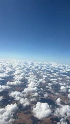 This is a video of a view from an airplane window, looking down at a vast expanse of fluffy white clouds spread across a blue sky. Below the clouds, the earth is visible, appearing as a patchwork of fields and possibly some buildings. The scene is brightly lit by the sun, casting shadows on the clouds and the ground. The camera is relatively stationary, providing a consistent aerial perspective. There are no people or explicit objects visible in the frame besides the natural elements of sky, clouds, and land. The video captures a serene and expansive aerial landscape.