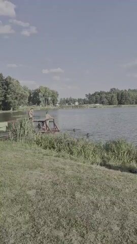 The video opens on a sunny day at a lake, with a clear blue sky and scattered clouds. The camera pans across the scene, revealing a lively beach area filled with people enjoying the water and the sand. In the foreground, a grassy embankment slopes down towards the lake, with tall reeds growing along the water's edge.

As the camera moves, we see a wooden pier with several individuals standing on it, some appearing to be preparing to jump into the water. Further out in the lake, other people are swimming and playing. The beach itself is crowded with sunbathers relaxing on towels and blankets, and children playing in the sand. Several beach umbrellas provide shade, and a few small structures are visible in the background.

The overall atmosphere is one of relaxation and recreation. The people appear to be enjoying a warm summer day at a popular swimming spot. The presence of beach umbrellas, towels, and swimwear indicates that this is a typical beach scene. The location cues suggest a lake or large pond in a rural or semi-rural area, with trees lining the distant shore. The time of day is clearly daytime, with bright sunlight casting shadows. No visible text is present in the video.