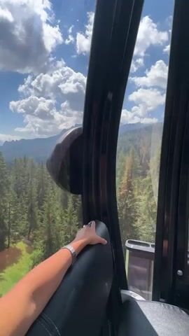 A first-person perspective video shows the interior of a gondola car as it travels up a mountain. The camera is positioned to show the view through the large windows. A person's arm with a watch is visible resting on a seat in the foreground. In the middle ground, a person with long dark hair is seated facing away from the camera, looking out the window. The gondola is ascending a forested mountain slope with a bright blue sky and white clouds overhead. Mountain peaks are visible in the distance. The interior of the gondola is dark and modern, with black seats and metallic accents. Safety instructions and a gondola number "33" are visible on the window. The video captures the scenic journey, emphasizing the natural beauty of the mountainous landscape.