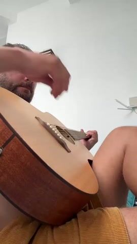 A man with his hair pulled into a bun is sitting down and playing an acoustic guitar. He is wearing shorts. The guitar is light brown and has a darker brown trim. The man is holding a pick in his right hand and strumming the strings. His left hand is on the fretboard. The background is a white wall with a light fixture on it.