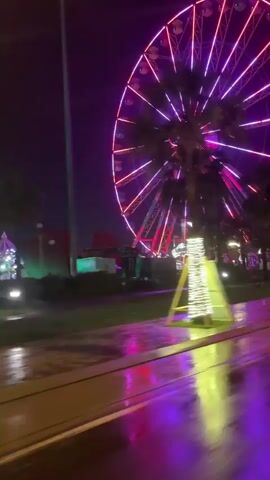 This media file depicts a vibrant night scene at an amusement park or fairground. The primary subject is a large, brightly lit Ferris wheel dominating the background, illuminated with a striking combination of purple and pink neon lights. The wheel has a circular logo at its center, which reads "The Chains" in white lettering.

In the foreground, the ground appears wet, suggesting recent rain or a damp environment, reflecting the colorful lights of the Ferris wheel and other park decorations. To the right of the Ferris wheel, a tall, triangular structure wrapped in bright yellow lights stands out against the dark sky. Further in the background, beyond the Ferris wheel, are glimpses of other park attractions, possibly including tents or buildings adorned with pink and red lights, and silhouetted trees. The scene is set at night, as evidenced by the darkness of the sky and the prominent use of artificial lighting.

The overall impression is one of festivity and excitement, with the glowing lights creating a magical atmosphere. The wet ground, however, might introduce a slightly melancholic or atmospheric mood, depending on interpretation. The location is specified as Tiến Thành, Vietnam. There are no visible people in this short clip.