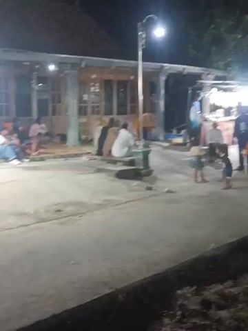 The video shows a nighttime scene of a street market. Several small stalls or kiosks are set up, displaying various goods, including what appear to be packaged snacks, drinks, and possibly some novelty items or toys. There are lights illuminating the stalls and the surrounding area. Several people are visible, some sitting or standing near the stalls, suggesting they are vendors or customers. One stall has a colorful display of lights. The ground is paved, and there are trees and buildings in the background, indicating an outdoor, possibly rural or semi-urban, setting. The overall impression is a bustling, albeit somewhat blurry due to low light and camera movement, evening market.