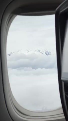 The video shows a view from an airplane window looking out at a snow-capped mountain peak rising above a sea of clouds. The window frame, typical of an airplane, is visible, as is a portion of an in-flight entertainment screen on the right side of the frame. The sky is overcast, and the overall color palette is muted, consisting primarily of whites, grays, and pale blues. There are no people or discernible actions visible in the video. The scene is static, focusing on the natural landscape viewed from a high altitude.