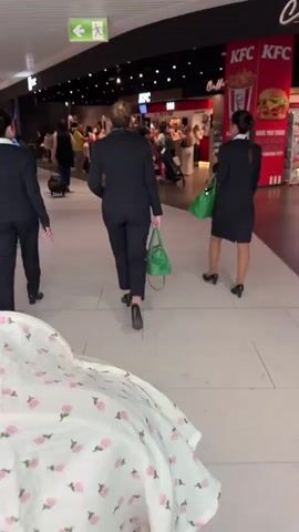 The video is shot from a low perspective, as if from a stroller. A white fabric with a pink floral pattern is visible in the foreground. As the camera moves forward, three women in matching navy blue suits walk away from the camera in a line. One pulls a blue suitcase, another carries a green handbag, and the third has no visible luggage. They walk down a wide corridor with a light gray tiled floor and a white ceiling with rows of fluorescent lights. Other people are seen walking in both directions. In the background, a KFC restaurant is visible, with a crowd of people near the counter. A green exit sign is overhead. The audio includes the sound of a baby crying.