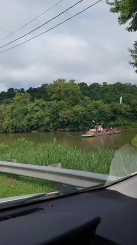 A wide shot shows a view from inside a vehicle looking out at a river. The river is wide and calm, with lush green trees lining the far bank. In the middle of the river, a tugboat is pushing a barge. The tugboat is red and white, and the barge is dark gray. There are power lines running across the top of the frame. On the near bank of the river, there is a metal guardrail, tall grass, and a wooden utility pole with several signs attached to it. One sign has directional arrows pointing to "NICHOLASVILLE" and "LEXINGTON." Another sign is a red octagonal "STOP" sign. The sky is overcast.