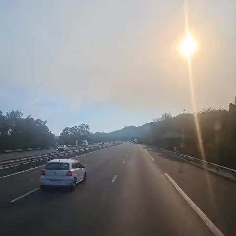 The video shows a view from inside a vehicle driving on a highway. The sun is shining brightly in the upper right corner of the frame, creating a lens flare effect. There are several cars on the road, with one white car in the foreground. Trees and hills are visible on either side of the highway. The sky is hazy with some clouds. The video appears to be taken during the daytime.