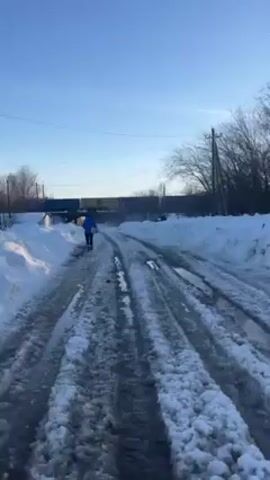 The video shows a person walking away from the camera on a snow-covered road on a bright, sunny day. The road is mostly clear of snow in the center, showing muddy tire tracks and puddles. Snowbanks line either side of the road. In the distance, there is a bridge or overpass with a dark structure on top. To the sides are trees, poles, and partially visible buildings. The sky is clear blue.