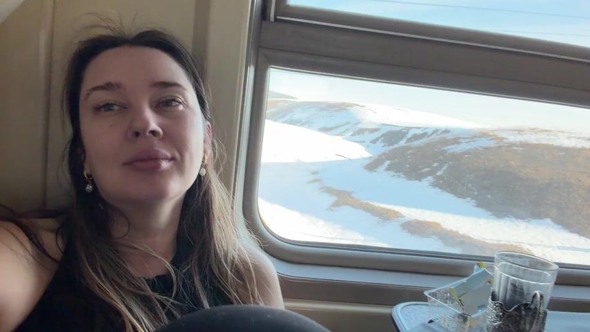 A woman is sitting on a train, looking directly at the camera with a slight smile. She has long brown hair and is wearing a black top. A train window is behind her, revealing a passing landscape of snow-covered hills and dry fields. There is a glass with a metal holder on a small table near the window. The train appears to be moving.