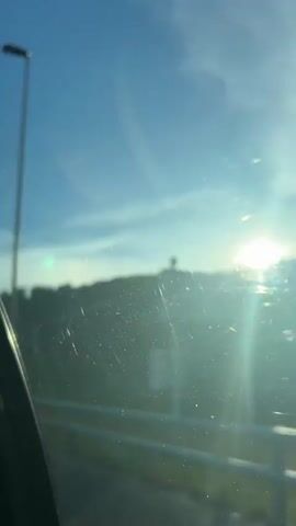 The video shows a view from a vehicle traveling on a road. A bright sun is visible in the upper right corner of the frame, casting a lens flare. The sky is blue with some wispy clouds. In the background, a dense forest covers a hillside. A metal guardrail runs along the side of the road in the foreground. The camera appears to be positioned to capture the scenery passing by.