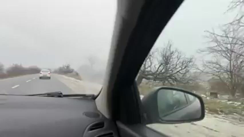 A view from inside a car driving on a road on a cloudy day. There is a white car ahead and some sparse trees alongside the road. The driver's side mirror is visible and reflects the scenery.