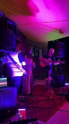 A group of people are performing music outdoors at night under a yellow canopy illuminated by pink and purple lights. A man on the right plays an acoustic guitar while standing on a stool. Two other people stand to his left, facing the camera. One person, wearing a puffer vest, holds a microphone. A laptop and audio equipment are visible on the left. The scene appears to be set up on a wooden deck or patio, with a red rug on the ground.