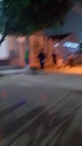 The video appears to be taken from a moving vehicle, possibly a motorcycle, at night.  The scene is an urban or suburban street with buildings and trees visible along the side. There are several people standing near a gate or fence in the foreground. One person is wearing dark clothing and white shoes, and appears to be holding something in their hand.  The video is shaky due to the movement of the vehicle. The street is lit by streetlights and lights from nearby buildings. There is background noise with music and voices.