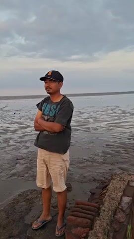 A man stands with his arms crossed, looking out at a muddy, wet landscape under a cloudy sky. He is wearing a dark t-shirt, light-colored shorts, a baseball cap, and flip-flops. In the background, there is a vast expanse of wet mud and a body of water. The sky is overcast with some lighter patches, suggesting it may be dawn or dusk. There are some rudimentary structures in the distance. The video pans across the landscape, showing the muddy terrain and the horizon.