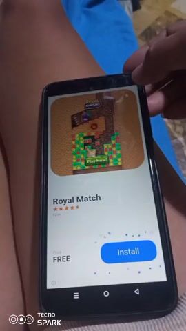 The video displays a smartphone with an advertisement for the game Royal Match. The screen shows the game's icon, title, star rating, and an "Install" button. The phone is resting on a person's lap, with a blue fabric visible in the background. The person's hand is partially visible at the top of the phone, holding it. The video is shot from a close-up perspective, focusing on the phone screen and its content. The advertisement is static, with no animation or interactive elements shown. The video appears to be a screen recording or a capture of a phone displaying an advertisement.