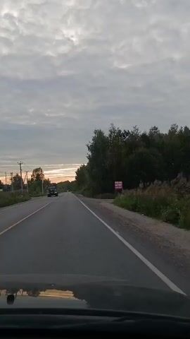 The video shows a view from inside a vehicle, traveling on a two-lane road. The sky is cloudy with a hint of sunset. On the left side of the road, there is a building, a grassy area, and utility poles with wires. On the right side of the road, there are trees, bushes, and a sign. A black car is parked on the left side of the road, and a dump truck is visible in the distance, driving ahead. The camera perspective suggests the video was recorded by someone in the passenger seat or from the dashboard.