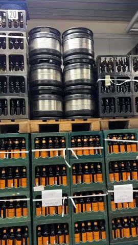 The video displays an indoor storage area or warehouse filled with beverages and packaging. In the foreground and midground, numerous green plastic crates are stacked on wooden pallets. These crates contain amber-colored bottles with orange labels and black caps, consistent with bottled beer. The crates are stacked approximately five to six high, with some stacks secured by white strapping or tape. Above these full crates, several stacks of dark gray or black empty plastic crates are visible, also on pallets, reaching a greater height. Some of these empty crates bear white numbers, such as "11" and "23". To the left of the frame, a tall stack of metallic, silver-colored beer kegs is positioned on a pallet, stacked about six high. The area is brightly lit, and no people or active processes are visible.