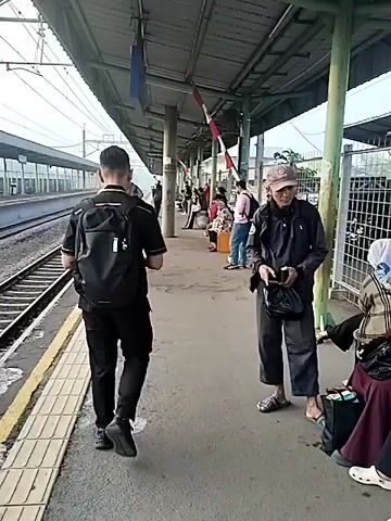 A video captured at a train station platform shows several people waiting. In the foreground, a person wearing a black shirt, black pants, and a black backpack walks away from the camera towards the train tracks. To the right of this person, another individual is seen from the waist up, wearing a dark jacket and a cap. They are holding a dark object in their hands, possibly a phone. Further down the platform, other individuals are visible, some seated, others standing. The platform itself is paved, with yellow tactile paving along the edge near the tracks. A covered waiting area with a roof supported by pillars is present. Train tracks run alongside the platform, with a white and red barrier gate visible in the distance. The environment appears to be outdoors, under a covered structure.