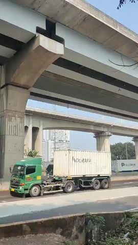 A white and red bus with a black and white logo travels right on a road. The bus is under an elevated highway. A large white shipping container with "INDORAMA" written on it is to the left of the bus. The shipping container is on a trailer with four axles and two wheels on each axle. The trailer is pulled by a yellow truck. A green covered truck is on the road to the right of the bus. Cars are visible in the distance to the right of the green covered truck. The environment is urban with a clear sky and some trees and brush at the bottom of the frame.