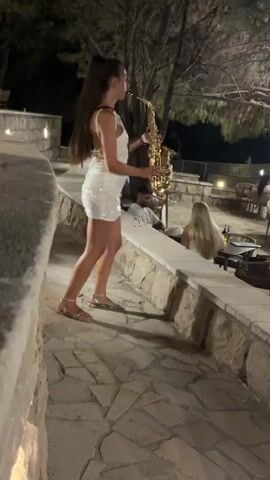 A young woman with long dark hair pulled into a ponytail, wearing a white sequined mini dress and silver sandals, plays a saxophone outdoors at night. She stands on a stone pathway in what appears to be an outdoor seating area of a restaurant or bar. The background shows tables and chairs, trees, and other patrons. The woman is in motion, swaying slightly as she plays.