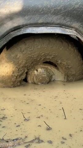 The video shows a dark grey SUV stuck in a muddy area. The front and rear wheels on the driver's side are submerged in thick, light-brown mud. The car body is also splattered with mud. The ground is completely covered in a layer of muddy water with small sticks scattered around. The video is taken from a close-up, side-view perspective.