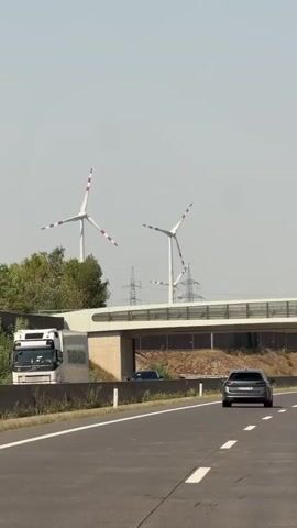 A perspective shot from inside a moving vehicle on a highway. The video captures a highway overpass with a truck visible on the left, and several cars on the highway. In the background, three wind turbines are visible, with two in the foreground appearing to be the main focus. The sky is clear and the environment seems to be a rural or semi-rural area, possibly in Europe given the design of the wind turbines.