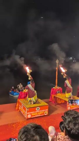 A group of people are gathered at night, possibly for a religious ceremony. In the foreground, several individuals are visible, some holding up their phones to record the event. In the background, on a raised platform, four individuals dressed in maroon and yellow attire are performing a ritual involving large flames. They are holding up ornate brass instruments with flames emerging from them. Smoke billows into the dark sky, and lights from a distant city or settlement are visible across a body of water. The platforms are decorated with red fabric and yellow flowers. One person in the water appears to be in a small boat.