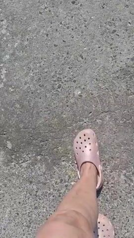 A first-person perspective video shows a person walking on a textured concrete surface. The person is wearing pink Crocs on both feet. The camera angle is looking down at the feet and the ground as the person walks. The concrete appears to be outdoors, with visible texture and some cracks. The lighting suggests it is daytime.