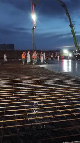 This video captures a construction scene at dusk or night. Several construction workers, some wearing reflective vests and hard hats, are actively engaged in pouring concrete. A concrete pump truck with a long boom is positioned in the upper left, directing the flow of concrete onto a reinforced subfloor made of rebar. The rebar grid is prominent in the foreground, partially covered by the wet concrete. The workers appear to be spreading and leveling the freshly poured concrete. In the background, other vehicles and lights suggest an active construction site. The sky is dark with some visible clouds.