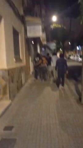 At night, an outdoor shot follows a group of approximately five to six individuals walking away from the camera on a sidewalk. The people are indistinct due to the distance and low light. To the left, a building features a light-colored lower facade and a darker upper section, with a closed window blind and a commercial sign mounted above. To the right, a tree with sparse leaves stands on the sidewalk, beyond which a street with parked cars is visible. A bright streetlight illuminates the scene from above. The camera moves forward, following the group.