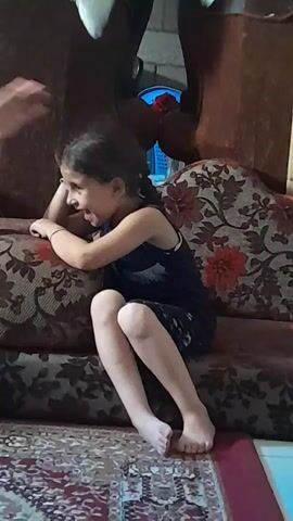 A young girl with brown hair in a ponytail is sitting on a patterned sofa. She is wearing a dark blue sleeveless dress. Her legs are bent, and her feet are bare. The girl is looking to her right and appears to be holding her hands up near her face. The sofa has a brown and floral pattern. A rug with a red and white pattern is visible on the floor in front of the sofa. The lighting suggests an indoor environment.