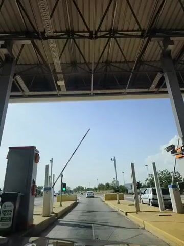 The video shows a view from inside a vehicle approaching a toll booth. The camera is looking forward through the windshield. The toll booth structure is overhead with metal beams and a roof. On the left side of the road, there is a grey booth with a green recycling bin. A white gate arm is raised, and a green traffic light is visible, indicating that the lane is open. Further ahead, cars are driving on the road. On the right side of the road, there are more toll booth structures and a white van is parked. The sky is clear and blue.