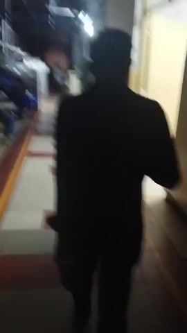 The video shows a person walking down a hallway. The person is silhouetted and appears to be wearing a dark shirt and pants. The hallway has a red floor with a checkered pattern. There are doors on either side of the hallway. In the background, there are other people and a sign. The lighting is dim.