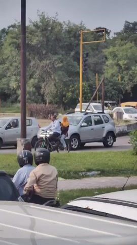 The video captures a street scene with multiple vehicles and people. In the foreground, a portion of a light-colored car is visible. Behind it, two individuals on a motorcycle, wearing helmets, are partially obscured by the car. The motorcycle rider is wearing a tan jacket and the passenger is wearing a light blue shirt. In the mid-ground, a silver SUV is stopped on the road. To the left of the SUV, a man is riding a bicycle, with a woman wearing a yellow hijab as a passenger behind him. The woman appears to be holding a mobile phone. Several other cars, including a white sedan, are also present on the road. The background consists of green trees and vegetation, with a traffic light visible on a yellow pole. The overall environment suggests an urban or suburban street with moderate traffic.