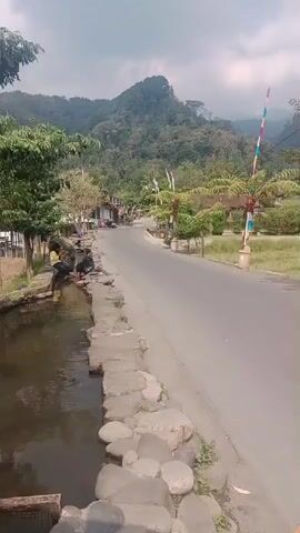 The video shows a scenic view of a road passing through a village with lush green mountains in the background. On the left side of the road, there is a narrow canal with stone lining and several people sitting by its edge. The people appear to be engaged in some activity near the water. The road is paved and curves into the distance, lined with trees and vegetation. Colorful flags are visible on poles along the roadside, adding a festive touch to the scene. The overall atmosphere is peaceful and serene, with a hint of local life and natural beauty.