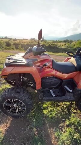 The video features a stationary orange ATV (All-Terrain Vehicle) parked in a scenic outdoor setting. The camera pans slowly from the left side of the ATV to its right, revealing the expansive landscape. The ATV has black seats and storage boxes, with "CFMOTO" and "CFORCE 800 XC" visible on its side, and "4x4" on the rear fender.

The setting appears to be a rural or wilderness area with a dirt path in the foreground, surrounded by green grass and scattered low vegetation. In the mid-ground, there are rolling hills or mountains. The sky is bright blue with scattered white, puffy clouds. The sun is low in the sky, creating a bright glow and lens flare, suggesting late afternoon or early evening, possibly sunset. The overall atmosphere is peaceful and natural. The location context is Anevo, Bulgaria.
