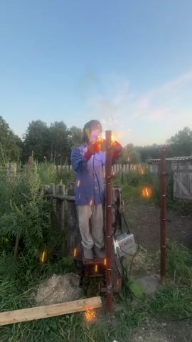 A person wearing a blue shirt, tan pants, red gloves, and a welding mask stands on a stool and welds a metal post in an outdoor environment. Sparks fly from the welding torch. A welding machine is visible on the ground next to the post. The background consists of greenery, trees, and a wooden fence. The sky is clear and blue, suggesting daylight.
