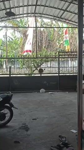 The video opens with a view of an outdoor covered area, possibly a carport or porch. In the foreground, the rear wheel and part of the frame of a black motorcycle are visible on the left. The ground is concrete and appears somewhat dirty, with some scattered debris. A metal pole runs vertically through the center of the frame. Beyond the covered area, through a metal gate and fence, is an outdoor scene with lush green vegetation, including what appears to be banana trees. Two flags are hanging from poles outside the fence; one is predominantly white with red accents, and the other is red, white, and green. The gate is ornate, made of black metal with decorative scrollwork. Behind the gate, there are more trees and foliage. In the middle ground, within the covered area but behind the gate, are several items including cardboard boxes, a red bucket, and a yellow bucket. Further to the right, inside the covered area, a large, dark, flat object is leaning against a wall, and a blue tube-like object is lying on the ground. A red object, possibly a small engine or pump, is also visible. In the lower right corner of the frame, there is a decorative item resembling a pumpkin or gourd, and a pair of flip-flops. The overall impression is of a somewhat cluttered but functional outdoor space, possibly in a tropical or subtropical climate.