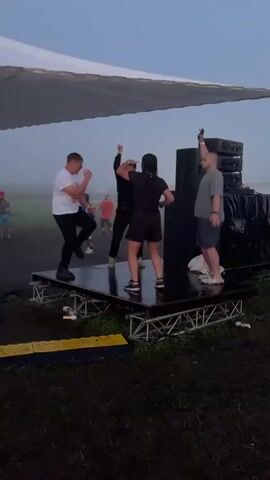 At the 0:00 mark, four individuals are dancing on a raised platform under a white canopy. The platform is supported by a metal truss structure. To the right of the platform are stacked black speakers. The environment appears to be an outdoor field with some fog present. The sky is overcast and light. In the background, there are other figures standing around, some appearing to be spectators. The lighting in the scene is dim.

At the 0:01 mark, the individuals continue to dance. One person in a white shirt and black pants is on the left side of the platform. A person in black clothing is in the center. Another person in black is to the right of the center. A person in a gray shirt and blue shorts is on the right side of the platform.

At the 0:02 mark, yellow lights begin to illuminate the scene, projected onto the underside of the canopy and from the front of the platform. The dancers continue their movements.

At the 0:03 mark, the yellow lights intensify and become more prominent, casting strong beams. The dancers are actively engaged in their activity, with raised arms and energetic movements.

At the 0:04 mark, the lighting remains vibrant with the yellow spotlights. The dancers' poses and movements are varied, suggesting a dynamic dance routine or freeform dancing.

At the 0:05 mark, the scene is consistent with the previous moments, with the dancers continuing to move under the illuminated canopy and the presence of the sound equipment. The fog remains a visible element in the background.