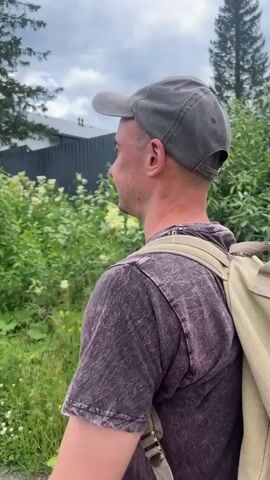 A man wearing a gray baseball cap and a dark t-shirt walks along a gravel road in a rural area. He carries a tan backpack. He turns to look at the camera and smiles. He then makes a funny face. The camera pans to show the road stretching into the distance, lined with trees and power lines. The video ends with a shot of a house under construction surrounded by greenery.