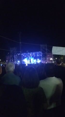 The media file depicts a live outdoor concert at night, likely in Rende, Italy. The scene is dominated by a brightly lit stage in the background, featuring a band performing. The stage is constructed with scaffolding and adorned with numerous lights, which are casting vibrant blue and yellow hues across the performers and the crowd.

In the foreground, a large audience is gathered, facing the stage. The audience members are mostly silhouetted against the stage lights, but some details are visible, such as the varying hairstyles and clothing of the individuals. The mood appears to be energetic and celebratory, as is typical of a concert atmosphere. The event is taking place after dark, as indicated by the black sky and the reliance on artificial lighting. There are no specific weather cues visible.

In the distance, beyond the stage, some urban lights can be seen, suggesting the concert is located in or near a populated area. There is also a large white banner or screen visible to the right of the stage, partially obscured by the crowd, but no legible text is discernible on it. The overall impression is one of a lively public gathering centered around a musical performance.