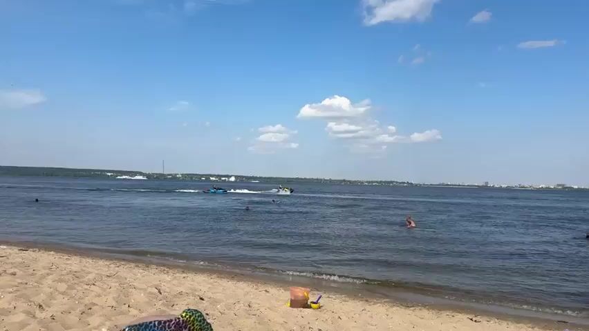 The video shows a wide body of water, likely a lake or river, under a bright, partly cloudy sky. The foreground features a sandy beach. In the water, several people are visible. Two individuals are swimming closer to the shore, with one person's head and shoulders above the water. Further out, two jet skis are in motion. One jet ski, colored blue and black, is traveling from left to right, leaving a wake. The second jet ski, primarily black with yellow accents, is also moving from left to right, generating a significant spray of water as it turns. The far bank of the water body is lined with trees and some buildings, suggesting a populated area in the distance. The scene depicts a summer day with recreational activities taking place on the water.