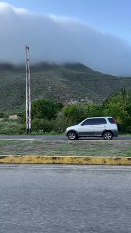The video shows a view from inside a vehicle traveling on a paved road. To the left, there is a yellow-striped curb and a median with some vegetation. Beyond that, a lush green landscape rises to a mountain shrouded in thick, white clouds. Palm trees and other dense foliage are visible in the mid-ground. A dark-colored SUV drives past in the opposite lane, its front end visible at the beginning of the clip and its rear end at around the 00:03 mark. Further in the distance, past the trees and the road, there are some structures that appear to be houses. The sky is a light blue with some white clouds. The overall impression is a scenic drive through a tropical or subtropical environment.