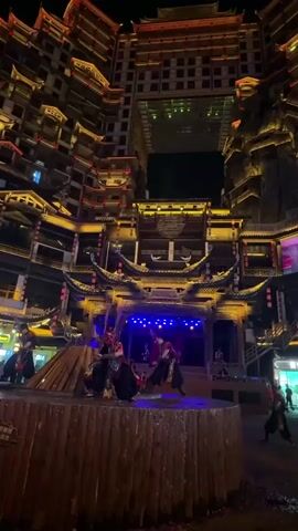 At night, several performers dressed in black, red, and silver traditional clothing are performing on a wooden stage. Each performer is holding a long thin sword. The stage sits in front of a building complex constructed in a traditional Chinese style, illuminated in yellow and blue lights. A rectangular gap runs through the center of the complex, revealing the dark night sky behind the structure.