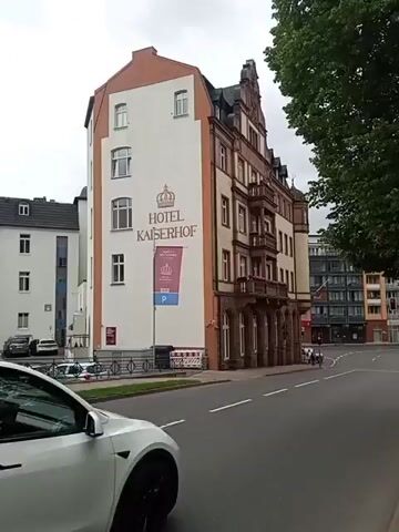 A white Tesla Model Y is driving down a street past a building with "HOTEL KAISERHOF" written on it. The hotel is a multi-story building with an ornate facade. There are trees and other buildings visible in the background. The sky is overcast.