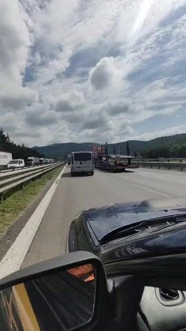 The video shows a view from inside a vehicle traveling on a highway. The perspective is from the driver's seat, looking forward and slightly to the right. A side mirror is visible in the lower left corner of the frame. The highway is busy with traffic, including trucks and a camper van. In the distance, there appears to be a traffic jam. The sky is cloudy with patches of blue sky and sunlight. The surrounding landscape consists of trees and hills. The time stamp indicates the video is from the early morning.